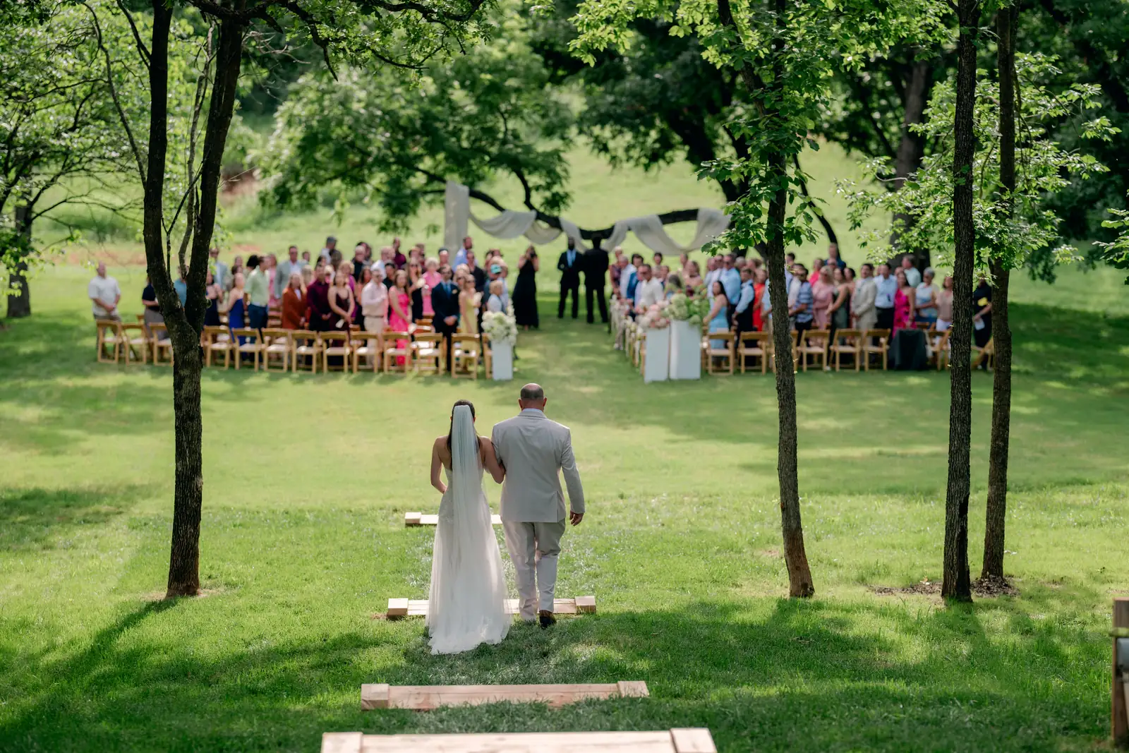 The bride walks the grass aisle toward the draped tree line