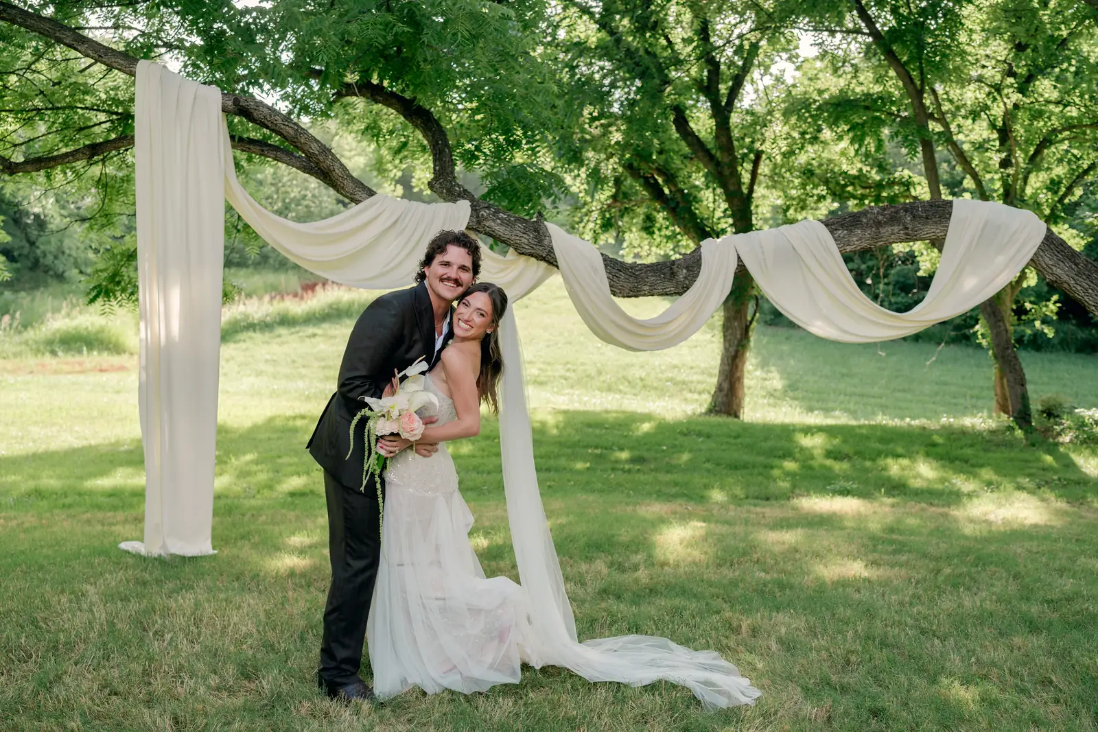 The couple laughing beneath ivory swags draped between the trees